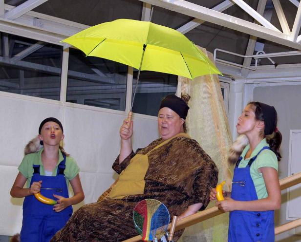 Eine Frau sitzt mit einem gelben Regenschirm, flankiert von zwei Mädchen mit Bananen in Overalls.