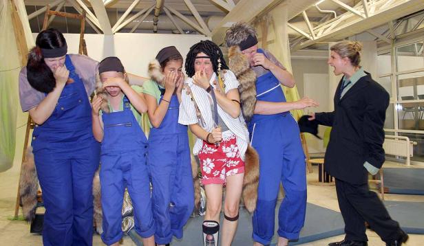 Eine Gruppe von Personen in Kostümen scheint sich die Nase zuzuhalten, während eine Frau im Anzug sie anschreit.