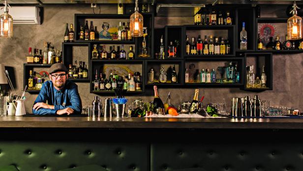 Ein Barkeeper steht hinter einer Bar, die mit Spirituosen und Cocktail-Zubehör gefüllt ist.