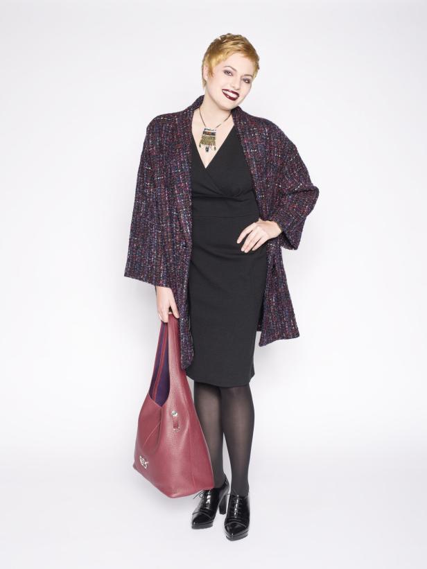 Eine Frau mit kurzem, blondem Haar trägt ein schwarzes Kleid, einen bunten Mantel und eine rote Handtasche.