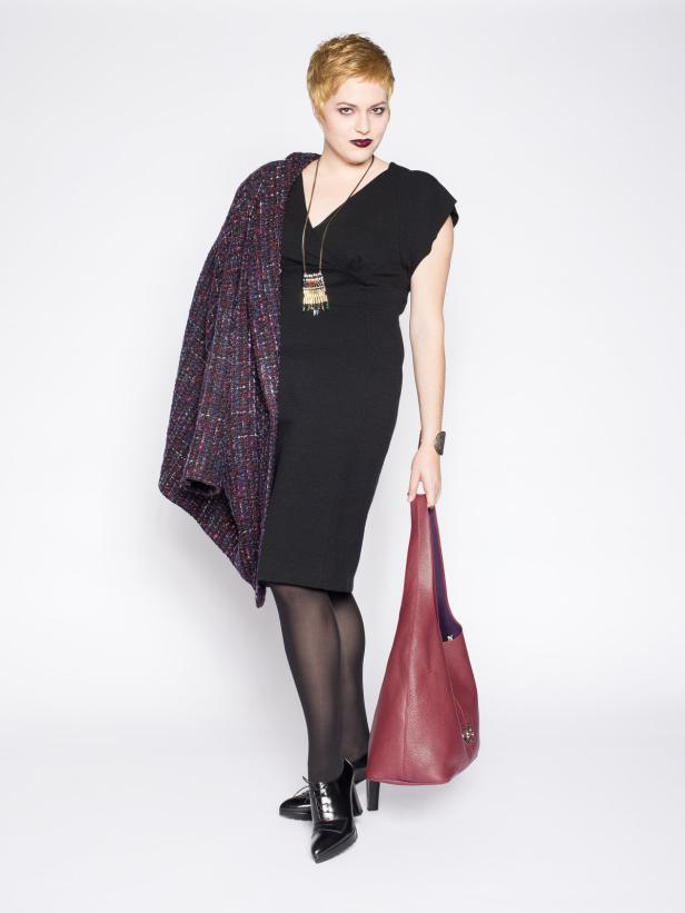 Eine Frau mit kurzem blonden Haar, schwarzem Kleid und roter Handtasche posiert vor einem weißen Hintergrund.