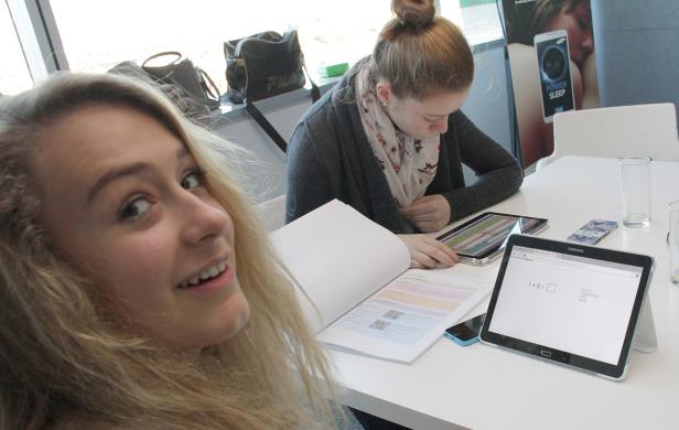 Zwei junge Frauen sitzen an einem Tisch und arbeiten mit Tablets und einem Buch.