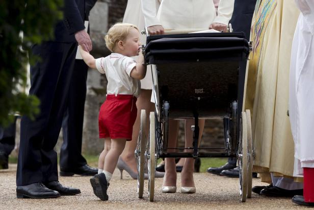 Ein kleiner Junge in roter Hose steht neben einem Kinderwagen.