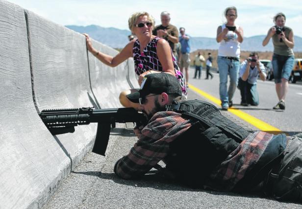 Ein Mann liegt mit einem Gewehr auf einer Autobahn, während andere Leute Fotos machen.