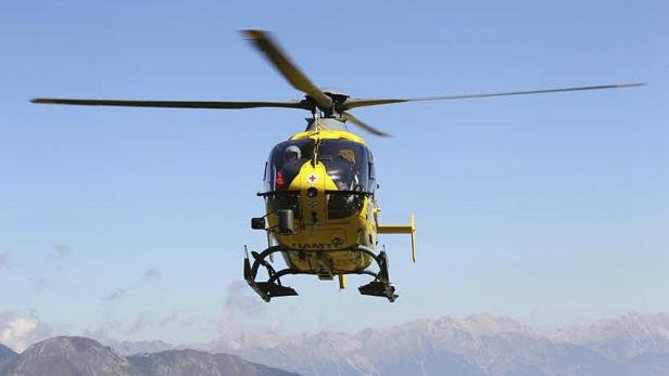 Ein gelber Rettungshubschrauber der DRF Luftrettung fliegt vor einer Bergkulisse.