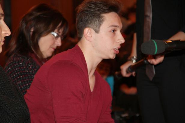 Ein junger Mann spricht in ein Mikrofon, während eine Frau im Hintergrund zusieht.