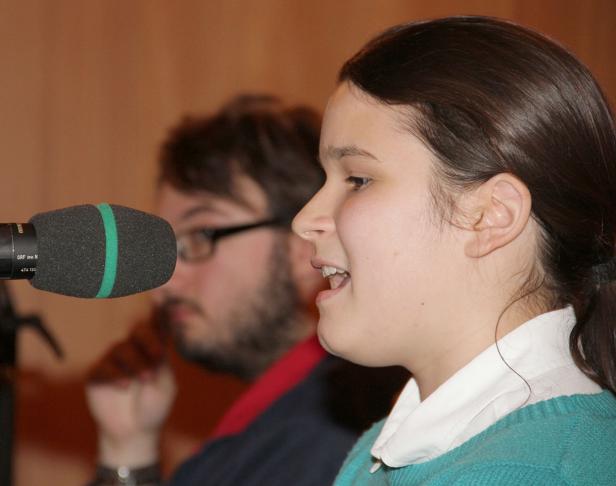 Eine junge Frau spricht in ein Mikrofon, während ein Mann im Hintergrund sitzt.