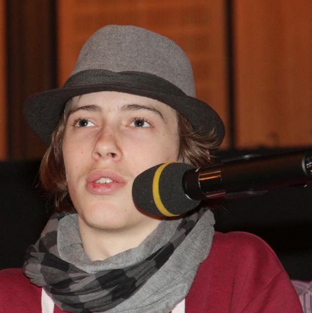 Ein junger Mann mit Hut und Schal spricht in ein Mikrofon.