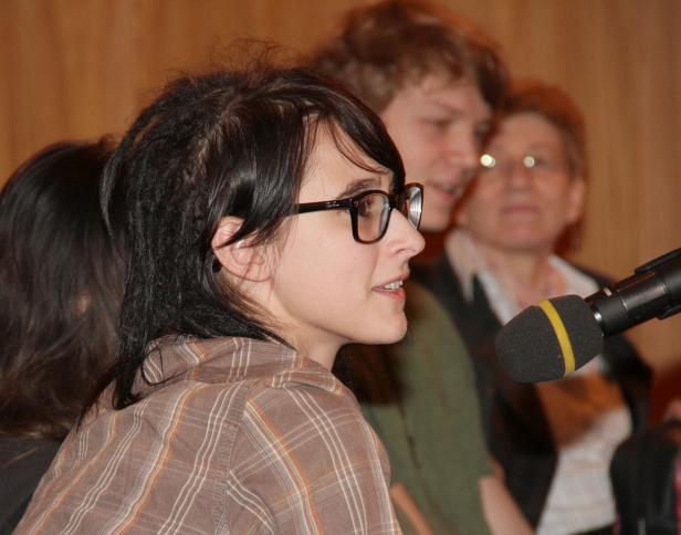 Eine junge Frau spricht in ein Mikrofon, im Hintergrund weitere Personen.