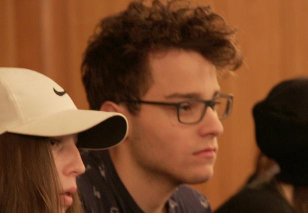 Ein junger Mann mit Brille und lockigem Haar sitzt neben einem Mädchen mit einer weißen Kappe.