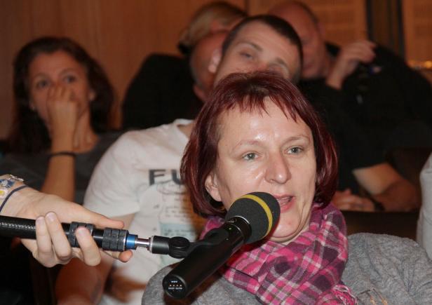 Eine Frau mit kariertem Schal spricht in ein Mikrofon vor Publikum.