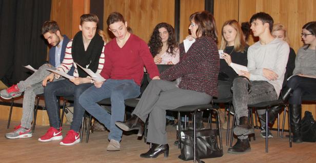 Eine Gruppe junger Leute und eine Frau sitzen auf Stühlen und halten Papiere in den Händen.