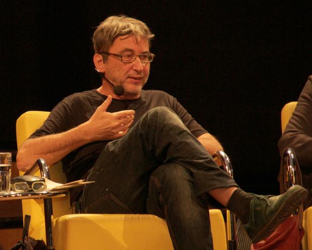 Ein Mann mit Brille und Mikrofon sitzt auf einem gelben Sessel und gestikuliert.