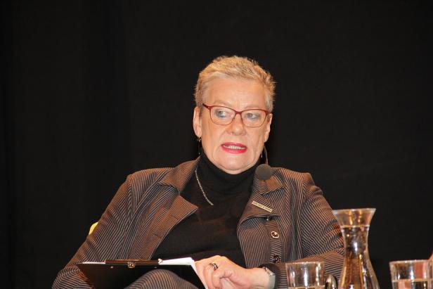 Eine Frau mit Brille und Mikrofon sitzt vor einem schwarzen Hintergrund.