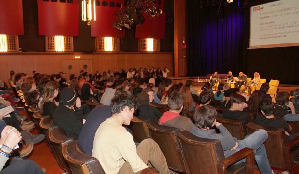 Publikum sitzt in einem Hörsaal und verfolgt eine Podiumsdiskussion.