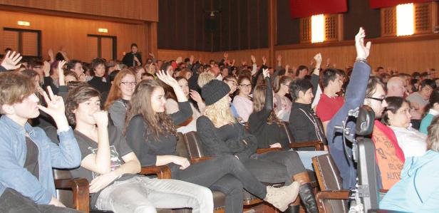 Ein Publikum sitzt in einem Auditorium und hebt die Hände.