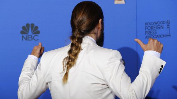 Ein Mann mit Zopf und weißem Anzug vor einem blauen Hintergrund mit Logos.