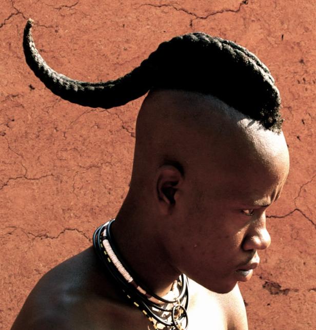 Ein junger Mann mit traditioneller Frisur und Halskette vor einer roten Wand.