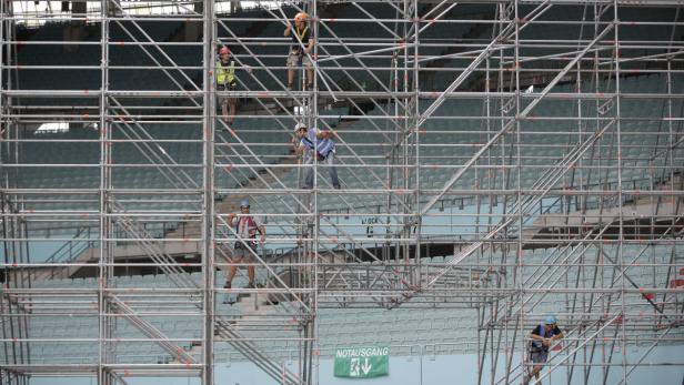 Mehrere Bauarbeiter montieren ein Gerüst vor den Sitzplätzen eines Stadions.