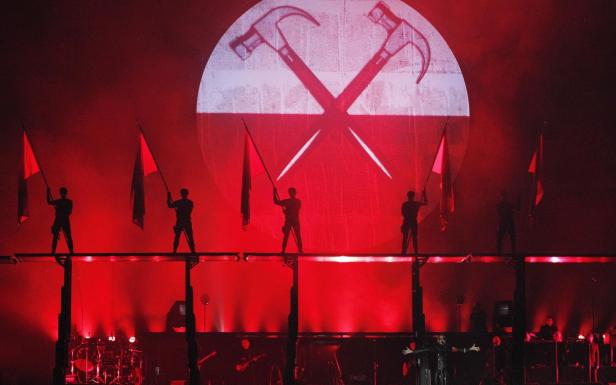 Eine Bühnenperformance mit Personen, die rote Flaggen vor einem Hammer-Symbol schwenken.