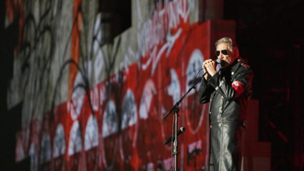 Roger Waters tritt in einer schwarzen Uniform mit einer Armbinde vor einer Projektion auf.