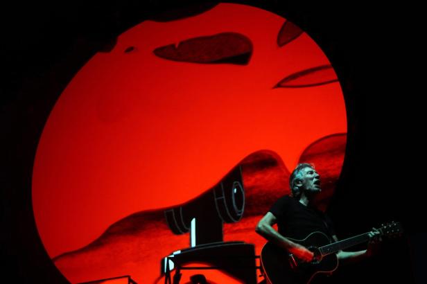 Roger Waters spielt Gitarre vor einer roten Leinwand mit einer Projektion.