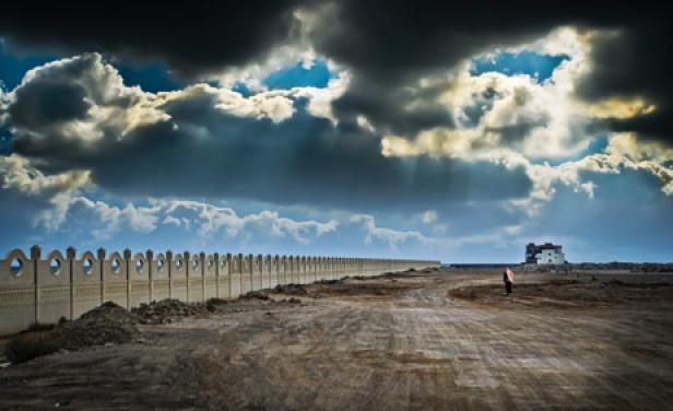 Eine Person steht auf einem Feldweg in der Nähe einer langen Mauer unter einem bewölkten Himmel.