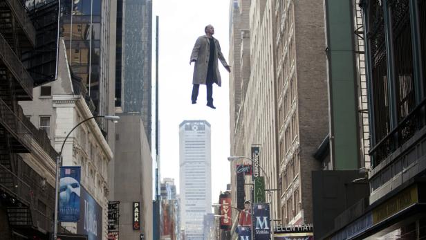 Ein Mann schwebt in einem Trenchcoat über einer Straße in der Stadt.