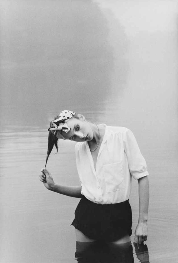 Eine junge Frau steht in einem Gewässer und hält eine Haarsträhne fest.