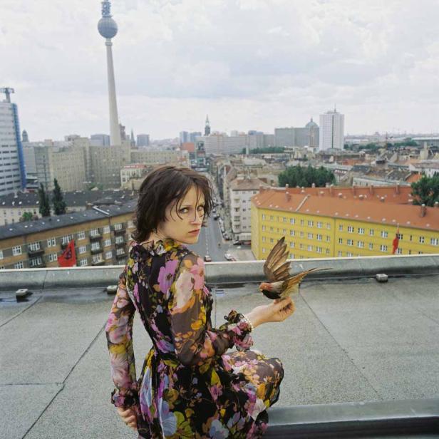 Eine Frau mit Blumenkleid hält einen Vogel auf einem Dach in Berlin.