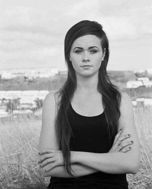 Eine junge Frau mit Sommersprossen steht mit verschränkten Armen vor einer Stadtlandschaft.