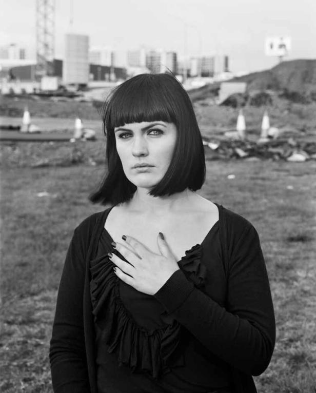 Eine Frau mit dunklen Haaren und dunkler Kleidung legt die Hand auf ihre Brust.