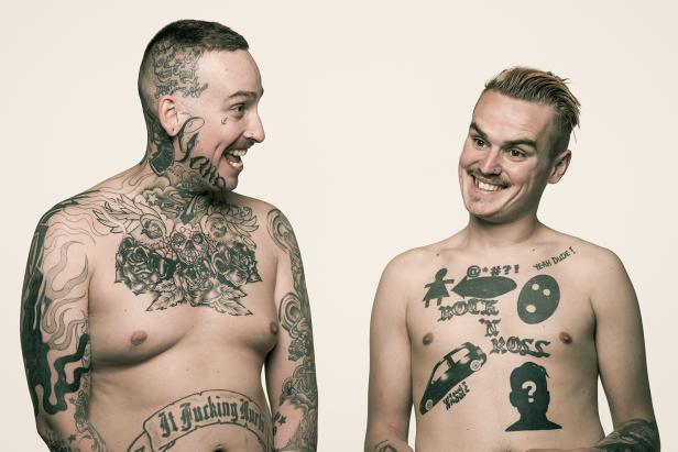 Zwei Männer mit Tätowierungen auf ihren Oberkörpern stehen sich gegenüber und lächeln.