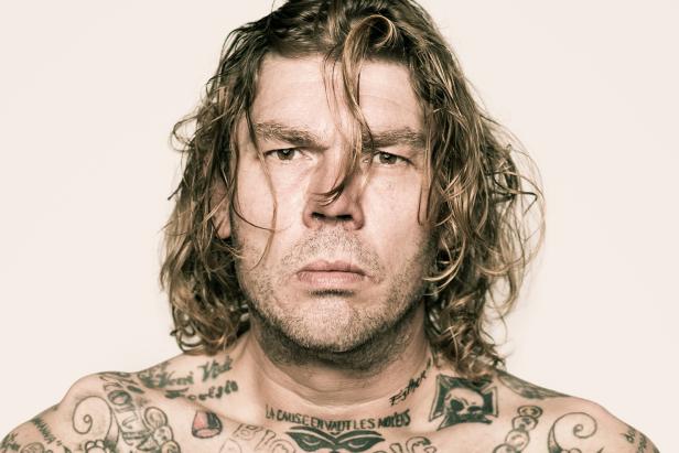 Ein Mann mit Tätowierungen und langen, gewellten Haaren blickt ernst in die Kamera.
