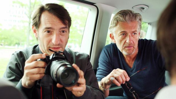 Zwei Männer sitzen in einem Auto, einer mit einer Kamera in der Hand.
