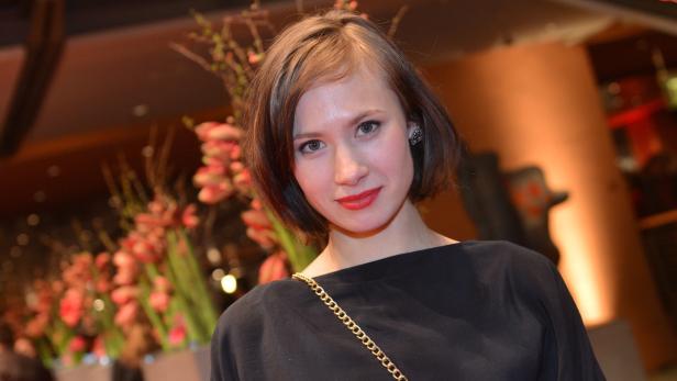Porträt einer Frau mit dunklem Oberteil und roten Lippenstift vor floralem Hintergrund.