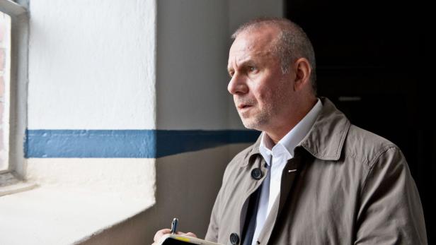 Ein Mann in einem Trenchcoat steht am Fenster und macht Notizen.
