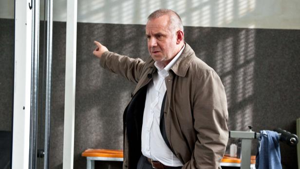 Ein Mann in Jacke und Hemd zeigt in einem Fitnessstudio nach links.
