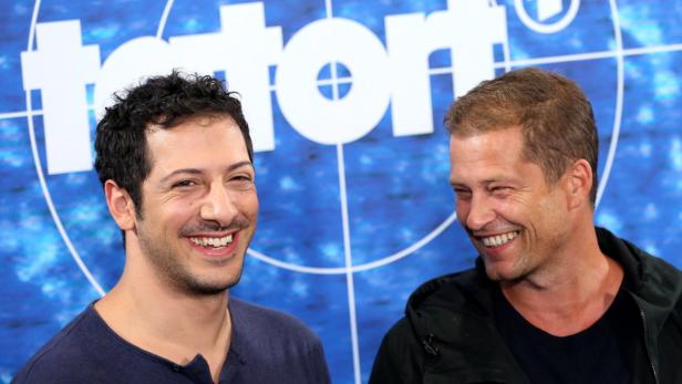 Die Schauspieler Elyas M'Barek und Til Schweiger lächeln vor einem blauen Hintergrund.