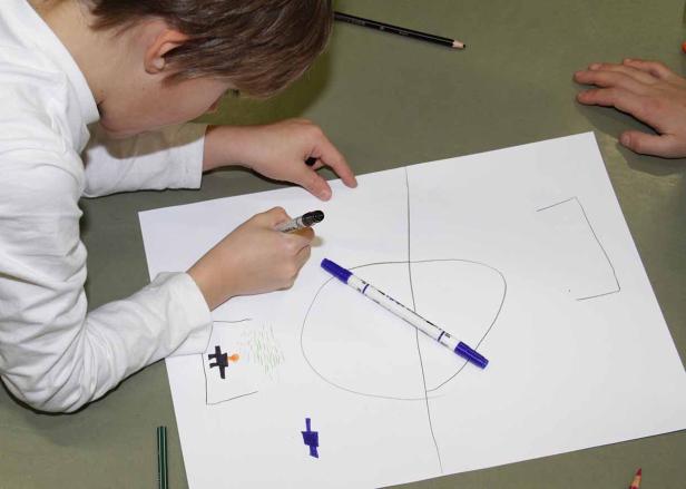 Ein Kind zeichnet mit Stiften auf ein Blatt Papier einen Fußballplatz.