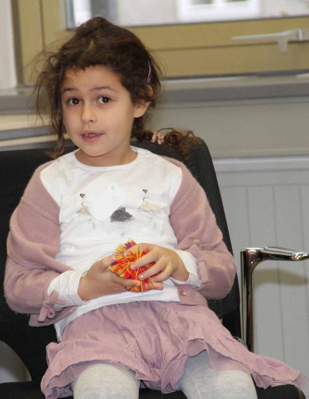 Ein junges Mädchen sitzt auf einem Stuhl und hält einen bunten Pompon in den Händen.