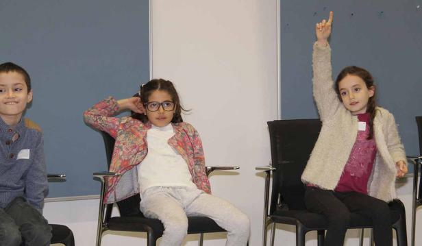 Drei Kinder sitzen auf Stühlen, ein Mädchen hat die Hand gehoben.