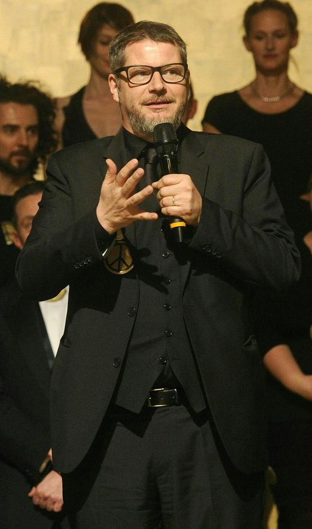 Ein Mann mit Brille und Anzug spricht in ein Mikrofon, eine Friedensikone tragend.