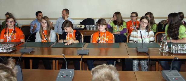 Eine Gruppe Jugendlicher sitzt an einem Tisch mit Mikrofonen und trägt T-Shirts mit der Aufschrift „World Peace“.