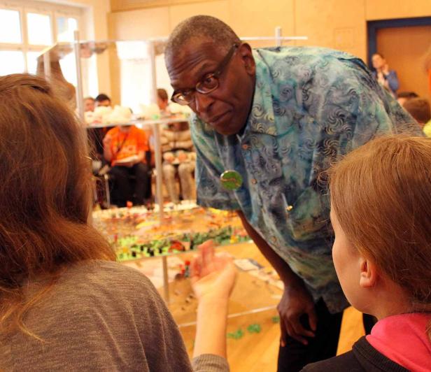 Ein Mann mit Brille beugt sich über ein Modell und spricht mit zwei jungen Frauen.