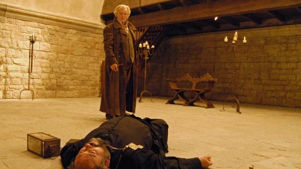 Ein Mann steht über einem leblosen Priester in einem Steingebäude.