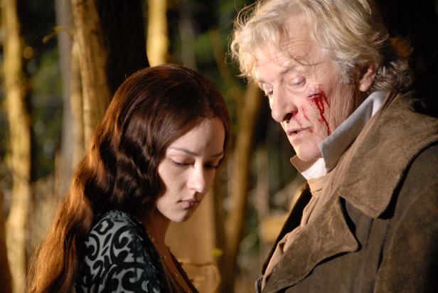 Eine junge Frau und ein älterer, verletzter Mann stehen im Wald.