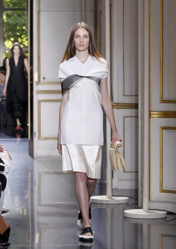 Eine Frau auf dem Laufsteg präsentiert ein weißes Kleid mit schwarzem Detail und eine Clutch.