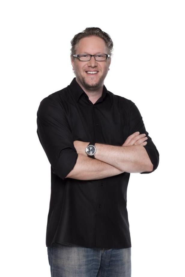 Ein Mann mit Brille, schwarzem Hemd und Jeans steht mit verschränkten Armen vor einem weißen Hintergrund.