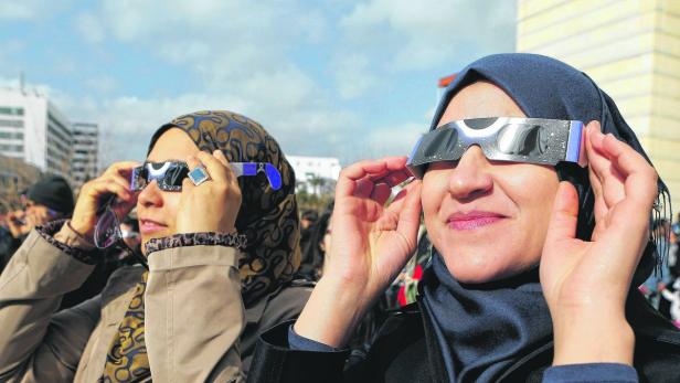 Zwei Frauen mit Kopftüchern beobachten mit speziellen Brillen eine Sonnenfinsternis.